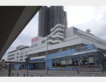 Straßenansicht eines großflächigen, mehrstöckigen Gebäudes mit weißer Fassadengestaltung und geometrischen Formen, davor ein gläserner Eingangsbereich mit Friseursalon. Im Hintergrund ein hohes Gebäude, das mit Gerüsten versehen ist, sowie eine Straße mit Straßenschildern und Passanten.