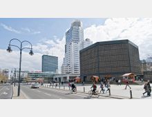 Straßenszene in einer Stadt mit modernen Hochhäusern im Hintergrund, darunter ein gläsernes Gebäude. Rechts im Bild steht die Kaiser-Wilhelm-Gedächtniskirche mit ihrem markanten kubischen Glockenturm. Fußgänger und Fahrradfahrer sind auf einem breiten Bürgersteig unterwegs, während links eine mehrspurige Straße verläuft.