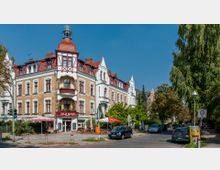 Ecke eines historischen Wohn- und Geschäftsgebäudes mit hellgelber Klinkerfassade, weißen Stuckverzierungen und roten Satteldächern; im Erdgeschoss befindet sich ein Restaurant mit Außensitzplätzen unter Sonnenschirmen. Im Vordergrund eine ruhige Straße mit geparkten Autos, umgeben von Bäumen und einem Straßenschild für eine Tempo-30-Zone.