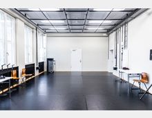 Heller, leerer Proben- oder Trainingsraum mit schwarzem Boden und weiß gestrichenen Wänden. Links befinden sich große Fenster und ein paar Stühle, rechts Kabelkanäle sowie ein Tisch; an der Stirnwand steht eine weiße Tür unter einer Rasterdecke mit rechteckigen Leuchten.