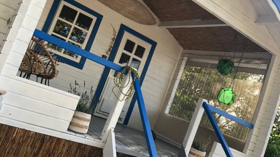 Kleines weißes Gartenhaus mit blauen Fensterrahmen und einem Vordach aus Holz, das mit einem Schilfgeflecht verkleidet ist. Eine Holztreppe mit blau-weißen Geländern führt zur überdachten Veranda, die mit Korbmöbeln und Pflanzen dekoriert ist.