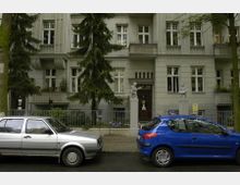Eine mehrstöckige Wohnhausfassade mit klassizistischen Elementen, einschließlich symmetrischer Fensteranordnung und kleiner Balkone, flankiert von zwei Bäumen. Vor dem Gebäude stehen zwei parkende Autos (ein silbernes und ein blaues) entlang der Straße.