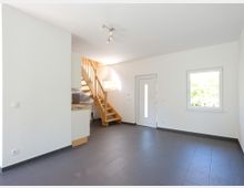 Heller Wohnraum mit weiß gestrichenen Wänden, grauem Fliesenboden und einer Holztreppe, die in ein höheres Stockwerk führt. Im Raum befinden sich ein Fenster mit Blick ins Grüne, eine weiße Eingangstür und eine kleine, teilweise sichtbare Küchenzeile.