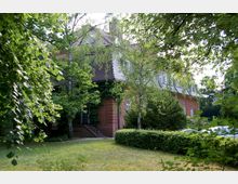 Backsteingebäude mit Mansardendach, teilweise von Bäumen und Sträuchern umgeben, im Vordergrund eine gepflegte Rasenfläche und Heckenbegrenzung.