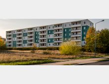 Wohnblock mit fünf Etagen in Plattenbauweise, grün-weiße Fassade, umgeben von Bäumen, Sträuchern und einer Wiese im Vordergrund, bei Tageslicht aufgenommen. Ein Gehweg und eine Straßenlaterne sind rechts im Bild zu sehen.