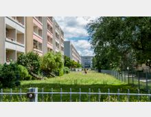 Mehrstöckige Wohngebäude mit Balkonen flankieren eine Grünfläche mit Bäumen, Sträuchern und einem umzäunten Bereich, in dem Wäsche aufgehängt ist. Der Himmel ist teils bewölkt, und der Bereich wirkt von Gehwegen und Zäunen umgeben.