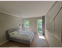 Ein minimalistisches Schlafzimmer mit einem grauen Bett und einer weißen Bettdecke, darüber ein aufgehängtes Moskitonetz. An der rechten Wand hängt ein großes Bild mit einer Hängebrücke, und es gibt zwei Fenster, durch die Tageslicht und grüne Bäume sichtbar sind. Der Boden besteht aus hellem Holzlaminat, ergänzt durch runde Teppiche in Weiß.