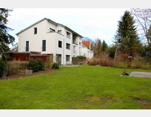 Einfamilienhaus mit weißer Fassade und Flachdach, umgeben von einem großen Garten mit Rasenfläche, spärlicher Bepflanzung und vereinzelten Bäumen. Im Hintergrund sind weitere Häuser und dichter Baumbestand zu sehen.