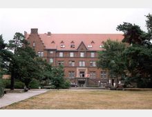 Ein historisches Backsteingebäude mit roten Ziegeldächern und zahlreichen Fenstern, umgeben von Bäumen und einer Rasenfläche. Im Vordergrund führt ein gepflasterter Weg durch eine parkähnliche Umgebung.