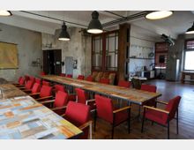 Ein großer, industriell gestalteter Besprechungsraum mit langen Holztischen und roten Stühlen, umgeben von rohen Wänden und dekorativen Elementen wie alten Lampen, Regalen und großen Fenstern. Im Hintergrund sind eine Holztür, eine historische Trennwand mit Glaseinsätzen und diverse Möbel zu sehen.