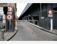 Einfahrt mit einer gesperrten Schranke vor einem industriellen Metalltor, flankiert von Verkehrsschildern, darunter ein Höhenbegrenzungszeichen mit "4 m", ein Gewichtsbegrenzungszeichen mit "8" und ein Durchfahrtsverbotsschild mit dem Zusatz "Anlieger frei". Rechts daneben verläuft eine massive Stahlkonstruktion.