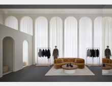 Moderner Showroom mit hohen, weißen Vorhängen, bogenförmigen Durchgängen und zeitgenössischem Design. Braunes Samtsofa, runder Holztisch und Kleiderständer mit Kleidungsstücken sowie Schaufensterpuppen definieren den Raum.