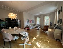 Großzügiges modernes Wohnzimmer mit Essbereich. Der Raum hat hohe Decken mit Stuckelementen, Parkettboden, einen runden Marmortisch mit gepolsterten Stühlen, eine Küchenzeile mit schwarzen Schränken und ein Sitzbereich mit Sofa und Sesseln vor großen Fenstern mit Vorhängen.