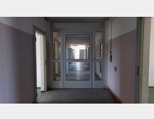 Ein schmaler Flur in einem Gebäude mit abgenutzten, weiß-rosafarbenen Wänden und einem gefliesten Boden, der zu einer Glastür führt. Hinter der Tür ist ein weiterer beleuchteter Gang sichtbar, mit offenen Türen auf beiden Seiten.