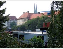 Stadtansicht mit einem historischen Gebäude mit rotem Ziegeldach und zwei Kirchtürmen im Hintergrund, umgeben von Bäumen und moderner Bebauung im Vordergrund, darunter ein Wohnmobil und dichter Bewuchs.