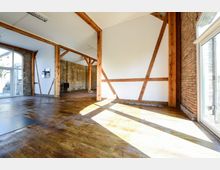 Großer, lichtdurchfluteter Raum mit Holzbalken, Backsteinwänden, großen Fenstern und Holzboden im Loftstil. Die Wände bestehen teils aus weiß gestrichenen Flächen, teils aus sichtbaren Backsteinen.