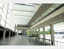 Weitläufiger, moderner Innenbereich eines Gebäudes mit hohen Decken, großen Fenstern und viel Tageslicht. Glasgeländer und eine Rolltreppe prägen die Raumgestaltung, während Büros und Besprechungsräume auf der oberen Etage sichtbar sind.