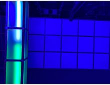 Ein Raum mit blauer Beleuchtung, dessen Hauptmerkmale eine Wand aus quadratischen, gleichmäßig angeordneten Paneelen und eine leuchtende, grün-blaue Säule aus transluzentem Material sind. An der Decke sind technische Geräte und Kabel sichtbar.