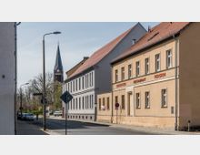 Straßenszene in einer Kleinstadt mit hellen Gebäuden, darunter ein gelbes Haus mit der Beschriftung "Biergarten", "Restaurant" und "Pension". Im Hintergrund ragt der Kirchturm einer Backsteinkirche mit spitzem grünen Dach empor, umgeben von Bäumen.