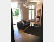 Wohnzimmer mit Holzboden, einer schwarzen Ledercouch, einem rechteckigen Glas-Couchtisch und einem braunen Teppich. Große Fenster mit Blick auf Grünanlagen lassen viel Tageslicht herein; ein Kronleuchter hängt an der Decke, und ein kleiner Holztisch steht in der Ecke.