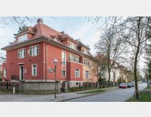 Ecke einer Wohnstraße mit einem zweistöckigen, roten Gebäude im Stil eines Stadthauses mit Satteldach und weißen Fensterrahmen. Im Vordergrund stehen eine Laterne, ein Straßenschild (Markischer Weg) und teils unbelaubte Bäume, während rechts geparkte Autos entlang der gepflasterten Straße zu sehen sind.