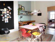 Wohnküche mit einem Tisch, der mit einer rot-weiß karierten Tischdecke bedeckt ist, umgeben von bunten Stühlen. An der Wand hängen ein schwarzes Memoboard mit Fotos und eine Regalfläche mit Büchern und Gläsern; eine hängende Lampe beleuchtet den Raum. Im Hintergrund ist ein weiterer Wohnbereich durch eine geöffnete Tür sichtbar.