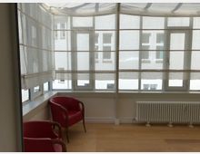 Ein heller Raum mit großen Fenstern und halbtransparenten Rollos, die das Tageslicht filtern. Im Vordergrund stehen zwei rote Sessel auf einem hellen Holzboden, vor einem Heizkörper an der Wand.