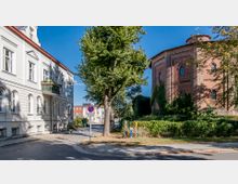 Straßenszene mit einem historischen, runden Backsteingebäude auf der rechten Seite, überwuchert von Efeu, und einem hellen, weißen Wohngebäude mit Balkonen links. Ein breiter Baum steht nahe der Straßenkreuzung, umgeben von Gehwegen und Pflastersteinen.