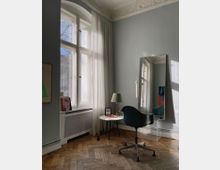 Heller Raum mit hohen Fenstern und weißen Vorhängen, ergänzt durch ein Wandspiegel. Der Boden ist aus Fischgrätparkett, und die Einrichtung besteht aus einem schwarzen Stuhl mit Rollen, einem runden Beistelltisch und einer Lampe.