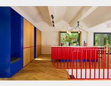 Innenraum mit modernem Design: Eine Küche mit auffälliger, rot-gestreifter Insel, blauen und holzfarbenen Wandschränken, einem Holzfußboden in Fischgrätmuster und großen Fenstern, die den Blick auf Bäume bieten. Eine rote Metalltreppe im Vordergrund und eine minimalistische Lampe an der Decke vervollständigen das Design.