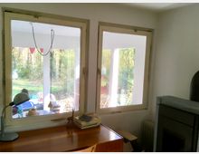 Innenraum mit einem Holztisch, auf dem Lampen, Bücher und Dekorationsgegenstände stehen, vor zwei großen Fenstern. Durch die Fenster sind eine überdachte Veranda sowie ein Garten mit Bäumen zu sehen.