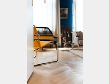 Ein modernes Wohnzimmer mit hellem Fischgrätenparkett, einem gelben Designerstuhl mit Metallrahmen und großen Fenstern mit weißen Vorhängen. Im Hintergrund sind eine blaue Wand, ein Bildrahmen und weitere Möbelstücke erkennbar.