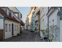 Schmale gepflasterte Gasse mit Reihen bunter historischer Gebäude, die pastellfarben gestrichen sind. Blumenkästen und Laternen schmücken die Häuserfassaden, während die Straße leicht gekurvt verläuft.