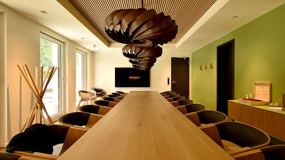 Moderner Konferenzraum mit langem Holztisch und vielen Stühlen, großen Fenstern an der linken Seite und einem Wandbildschirm am Ende des Raums. Auffällig sind die skulpturale, dunkle Pendelleuchte und die Holzlamellendecke; rechts eine grüne Akzentwand mit Türen und Sideboard.