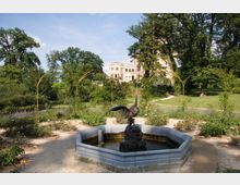 Ein Garten mit einem achteckigen Brunnen im Vordergrund, der eine Vogel-Skulptur zeigt, umgeben von Rosenbeeten und geschwungenen Rankhilfen. Im Hintergrund ist ein helles historisches Gebäude mit Türmen und Bögen, eingebettet in eine grüne, parkähnliche Landschaft mit großen Bäumen und geschwungenen Wegen, sichtbar.
