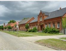 Straßenszene in einem Dorf mit historischen Backsteinhäusern, die an einer gepflasterten Straße liegen. Die Gebäude haben verziertes Mauerwerk und Satteldächer, im Vordergrund ein Verkehrsschild mit Geschwindigkeitsbegrenzung, umgeben von Bäumen und Rasenflächen.