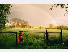 Ein weitläufiges Feld mit grünem Gras, im Hintergrund einige Bäume und ein leuchtender Regenbogen am Himmel. Im Vordergrund ein Metallzaun mit einem offenen Tor und ein Kind, das durch das Tor geht.
