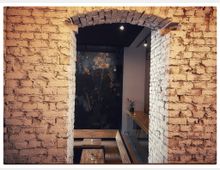 Innenraum mit freigelegter, weiß gestrichener Backsteinwand und einem bogenförmigen Durchgang. Dahinter befinden sich Holztische, eine Wand mit floralem Muster und vereinzelte dekorative Elemente wie eine Vase mit Blumen.