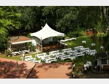 Gartenanlage mit Rasenfläche, auf der zahlreiche lange Tische und Bänke mit weißen Hussen in Reihen stehen. Im Hintergrund befindet sich eine kleine Bühne unter einem weißen Zeltdach, umgeben von Bäumen und kleinen Nebengebäuden.