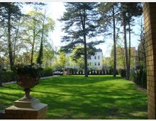 Grüne Parkfläche mit hohen Bäumen, umgeben von Wohnhäusern im Hintergrund. Im Vordergrund steht ein kunstvoller Blumenkübel auf einem gemauerten Sockel.