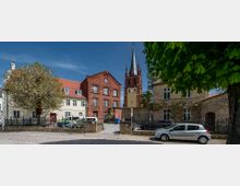 Eine Ansicht eines gepflasterten Platzes mit historischen Gebäuden, darunter eine rote Backsteinfassade, eine Kirche mit hoher Turmspitze und Uhr sowie gemischt hellere Wohnhäuser. Im Vordergrund sind Bäume, parkende Autos und ein Metallzaun zu sehen.