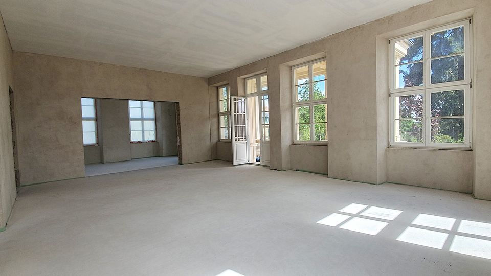 Unmöblierter, renovierungsbedürftiger Innenraum mit glatten, verputzten Wänden und hellem Boden. Mehrere große Sprossenfenster sowie eine Glastür lassen viel Tageslicht hinein und bieten Blick ins Grüne.
