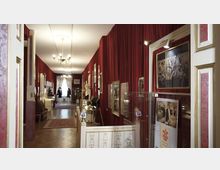 Langer, schmaler Museumsraum mit roten Wänden, an denen gerahmte Bilder und Poster hängen. Vitrinen mit Exponaten, ein roter Teppich auf dem Holzboden und ein Kronleuchter prägen den Raum; am Ende des Gangs sind Schaufensterpuppen mit historischen Kostümen zu sehen.