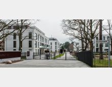 Moderne Wohnanlage mit mehrstöckigen, weißen Gebäuden, großen Fensterfronten und Balkonen, umgeben von Bäumen. Im Vordergrund ein Metalltor mit gepflastertem Zufahrtsweg und gepflegtem Grünstreifen.