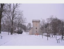 Historischer Steinbau mit Turm und Ziffernblatt, umgeben von schneebedeckten Wegen, Bäumen und Parkelementen in einer winterlichen Landschaft. Im Hintergrund sind leicht bewaldete Hügel und vereinzelte Spaziergänger sichtbar.