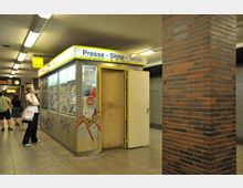 Kiosk in einer U-Bahn-Station mit der Aufschrift „Presse - Shop - Tabak“. Die Umgebung zeigt braune Fliesenwände, ein beleuchtetes U-Bahn-Schild mit der Aufschrift „Urbans“ und mehrere Personen im Hintergrund.