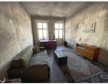 Langer, schmaler Wohnraum mit abgenutzten, teilweise abgeplatzten Wänden und zwei hohen Fenstern am Ende, die viel Tageslicht hereinlassen. Im Raum stehen ein altes Sofa, ein Schreibtisch mit Stuhl, ein großer Teppich, ein trunkähnlicher Couchtisch sowie ein Sideboard an der rechten Wand.