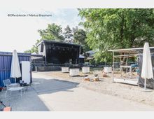 Außenbereich mit einer Open-Air-Bühne und Bühnequipment, umgeben von Bäumen. Im Vordergrund stehen weiße Gartenmöbel, Sonnenschirme und Tische, während leere Wassertanks und Holzpaletten im Gelände verteilt sind.