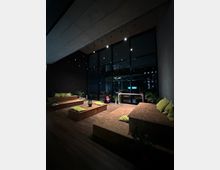 Moderner Innenraum mit minimalistischem Sitzbereich aus Holzpodesten, die mit gelben Kissen dekoriert sind. Der Bereich ist von großen Fenstern mit Blick auf eine nächtliche Stadtszene umgeben und wird von dezenten Deckenleuchten beleuchtet; Pflanzen sorgen für eine natürliche Atmosphäre.