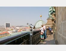 Personen stehen auf einer Aussichtsplattform an einer historischen Kuppel mit grüner Patina. Im Hintergrund erstreckt sich eine Stadtlandschaft mit modernen und historischen Gebäuden, ein Fernglas ist auf der Plattform montiert.